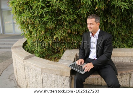 Businessman typing by laptop outside near green plant in   and looking at watch. Concept of ecology thinking for business. Successful man dressed in black suit working with computer outdoors.