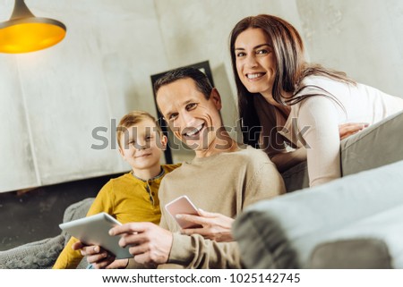 We love gadgets. Pleasant upbeat young family sitting on the couch and smiling at the camera brightly while posing with their gadgets