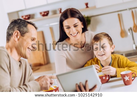 Having fun together. Pleasant young man showing the tablet to his family and watching the video together while they having breakfast in the morning