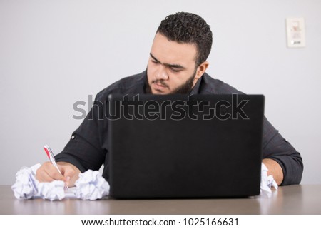 A frustrated employee sitting, trying to write something after cutting a lot of paper and balled it on the front of his laptop.