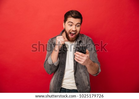 Picture of Happy screaming bearded man using smartphone and rejoices over red background