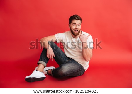 Picture of smiling bearded man sitting on floor isolated over red background wall. Looking camera pointing.