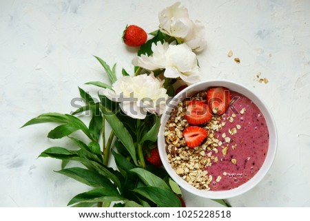 Smoothie bowl with granola and strawberry near white peonies on white background. Top view.