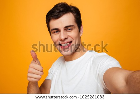 Portrait of a smiling young man showing thumbs up gesture while taking a selfie isolated over yellow background