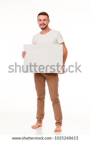 A young charismatic man with a white sign in his hands. You can use it to advertise, place a logo. He is dressed in brown pants, a white t-shirt, barefoot. Full length of the photo, different emotion