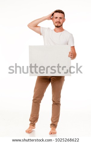 A young charismatic man with a white sign in his hands. You can use it to advertise, place a logo. He is dressed in brown pants, a white t-shirt, barefoot. Full length of the photo, different emotion