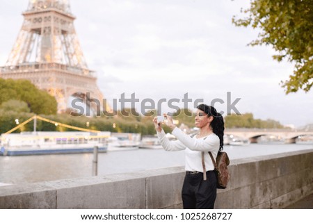 Student walking along embankment not far from Eiffel Tower and making photos of landmarks using smartphone. Young woman recently moved to Paris to live and study. Smiling black-haired female with