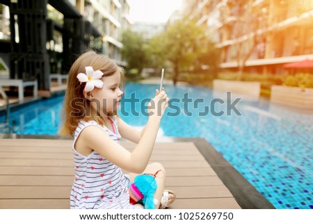 little kid girl take photo on table, tsitting close to blue swimming pool wearing flower and hold toy. concept of new technologies travel, recreation or holiday family time on sunny day