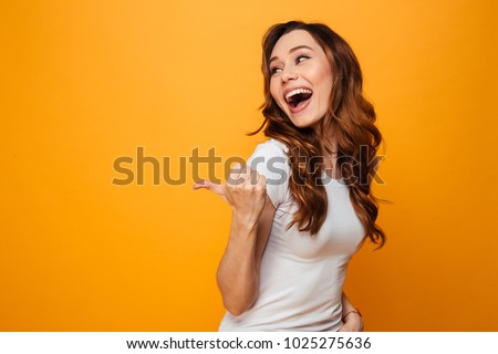 Laughing brunette woman in t-shirt looking and pointing away with thumb over yellow background