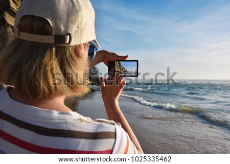 Woman in cap with a smartphone takes pictures of a beautiful sunset, ocean and rocky coastline on the portuguese beach.  Azenhas do Mar, Portugal