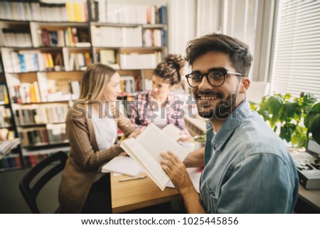 Portrait view of a nerdy handsome bearded male student in the library holding book while two female friends talk in the background. 