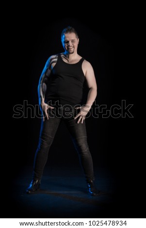 Young man in black T-shirt and blue jeans with bodypainting on shoulder and hand posing on black background with blue light
