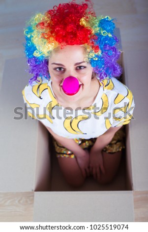Surprise! 1 April Fools' Day concept. Funny smiling girl in a clown suit sitting in a cardboard box and looking at the camera. Birthday.