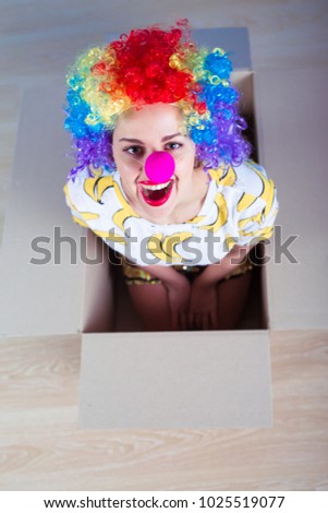 WOW! 1 April Fools' Day concept. Funny smiling girl in a clown suit sitting in a cardboard box and looking at the camera. Birthday.