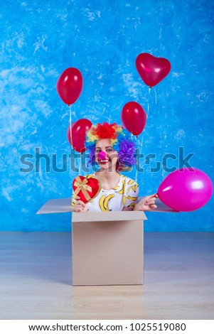 1 April Fools' Day concept. Funny laughing clown girl sits in a cardboard box with helium balloons and looking in camera. Birthday gift.