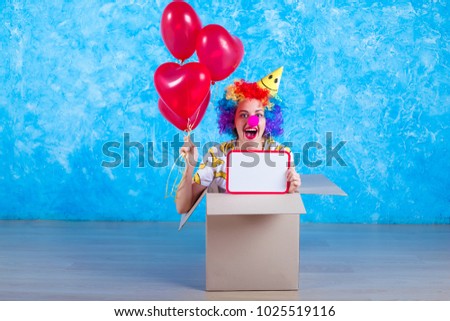 1 April Fools' Day concept. Funny laughing girl clown sits in a cardboard box with helium balloons and white board in hand. Birthday gift.