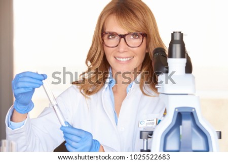 An executive middle aged medical assistant woman holding a test cube in her hand and preparing for medical lab test while sitting in front of microscope.