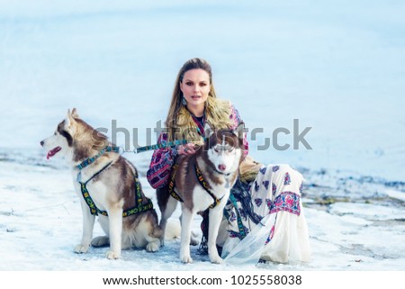 Beautiful blonde girl in a company of two  husky dogs sits near the winter river bank 