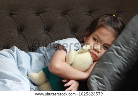4-5 year old Asian patient kid lie down on couch or sofa in patient suit with her doll.Skinny kid look sad,tired,pain and sick.Concept of childhood cancer awareness.