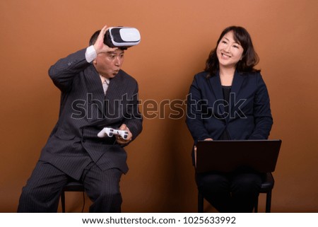 Studio shot of mature Asian businessman and mature Asian businesswoman together against brown background