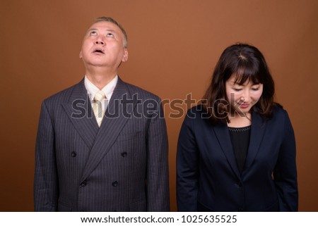 Studio shot of mature Asian businessman and mature Asian businesswoman together against brown background