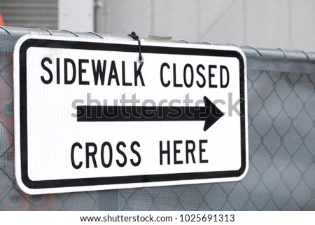 Close up on a Sidewalk Closed, Cross Here street sign, with a black arrow pointing right, on a chain link fence