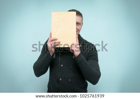 Happy man hides his face behind a wooden plaque plate in his hands and peeks out from there isolated on blue background.
