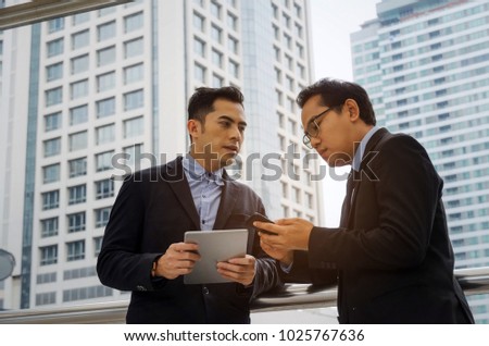 two business asian man talking and reading information about finance news in mobile phone tablet together standing in modern city, network technology, internet, successful, teamwork concept