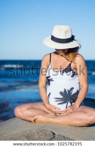 close up a pregnant woman in swimming suit and wears white hat, holding belly and sitting in beach, enjoy the sun in summer.
