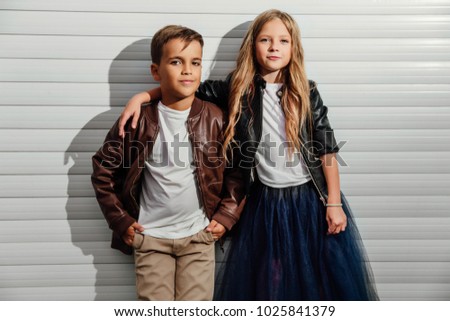 Portrait of two teenage school children on a garage door background in a city park street. Kids model friends, brown and black jackets, blue skirt, white T-shirt. Copy space for text message. Royalty-Free Stock Photo #1025841379