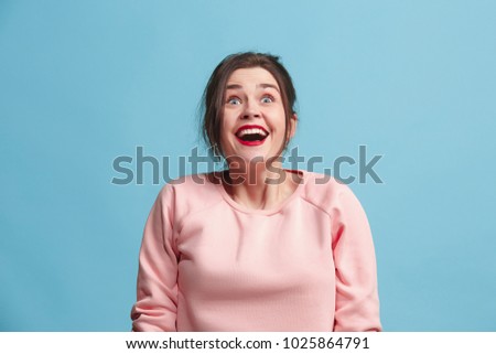 Beautiful female half-length portrait isolated on blue studio backgroud. The young emotional smiling and surprised woman standing and looking at camera.The human emotions, facial expression concept
