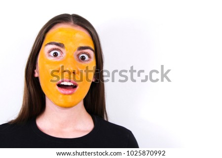 Young beautiful surprised girl with a mask of turmeric on her face on a white background. Face close-up with space for text