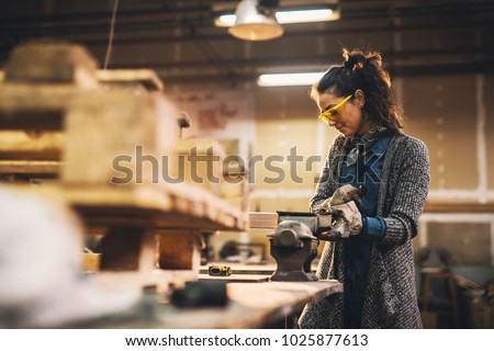 Portrait view of satisfied smiling middle aged professional female carpentry worker with steel vise on the table in the workshop. Royalty-Free Stock Photo #1025877613