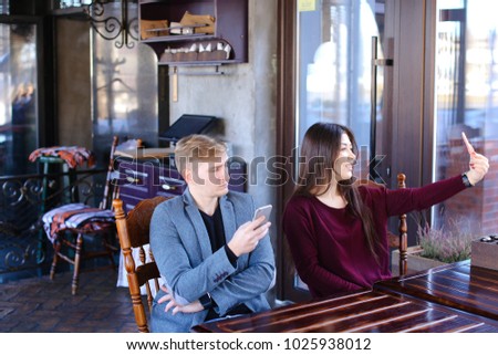 Physicist with smartwatch on hand in cafe with colleague taking selfies using smartphone wait friend, irritated guy checking social networks watching smiling Asian girl. Young people sitting near