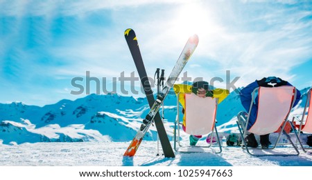 Image from back of vacationers in armchair, skis, sticks in snowy resort