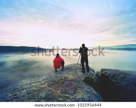 Photographer  with eye at viewfinder of camera on tripod stay on cliff and takes photos, talk friends. Autumn beautiful  misty landscape, misty sunrise  at horizon. Men in mountains