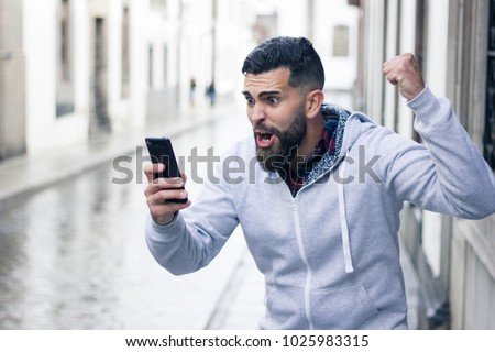 Enthusiastic young man looking at cellphone with victorious expression in the city. Overexcited hipster with grey hoodie holding mobile device and fist up. Online bet winner concept Royalty-Free Stock Photo #1025983315