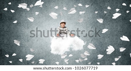 Young little boy keeping eyes closed and looking concentrated while meditating on flying cloud among flying paper planes with gray dark wall on background.