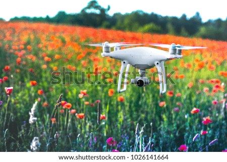 White quadrocopter, is flying high in the air, to take photos and record footage from above, in red poppy field. Drone with four motors, propellers, camera, warning lights. Romantic flowers background