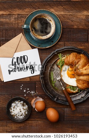 Croissant sandwich with fried egg and arugula, cup of coffee, greeting card "good morning", salt and eggshell on wooden background. Flat composition. Delicious and healthy breakfast