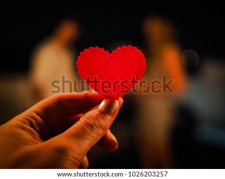 holding a red heart in front of a blurred couple