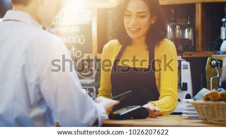 In the Cafe Beautiful Hispanic Woman Makes Takeaway Coffee For a Customer Who Pays by Contactless Mobile Phone to Credit Card System. Royalty-Free Stock Photo #1026207622