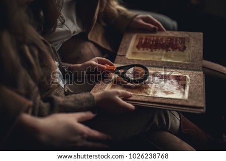 Child girl with woman in image of detective sits in armchair and looks old photo album with magnifier. Closeup.