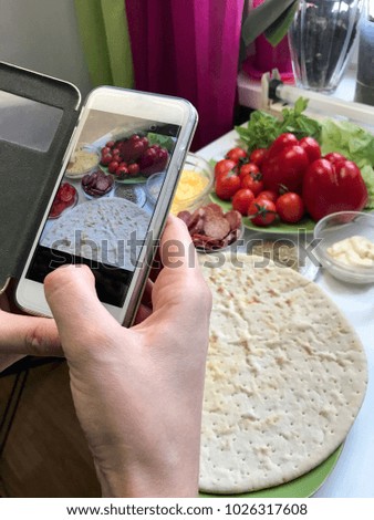 A girl takes pictures of a pizza on a smartphone. Necessary ingredients are on the table: cheese, sausage, pizza base and vegetables.