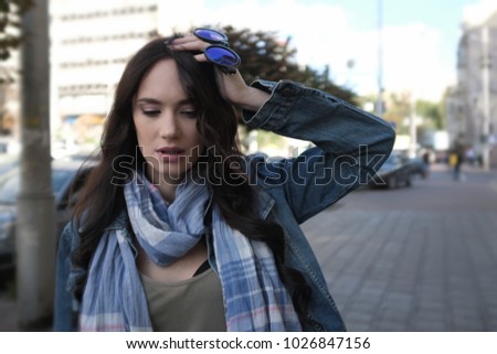 Pretty brunette girl disturbed is walking in a city street. She is wearing a blue jeans coat and blue scarf. Holding sunglasses in her left hand.