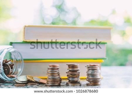 Pile of money coins outside the glass jar in front of books on blurred natural green background for financial and education concept