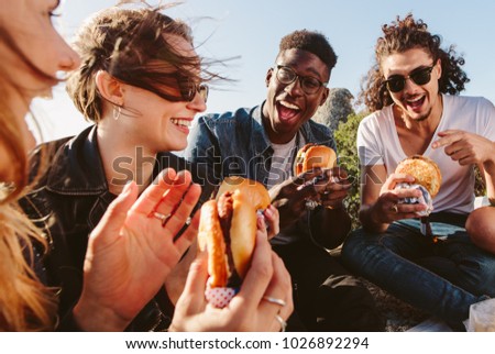 Group of friends sitting on mountain top eating burger. Excited young men and women enjoying and partying outdoors. Royalty-Free Stock Photo #1026892294