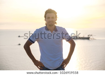 Handsome man brunette in a light t-shirt with his hands on the belt against the seaside background.