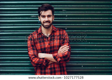 Young smiling man, model of fashion, wearing a plaid shirt with a green blind behind him. Guy with beard and modern hairstyle in urban background.