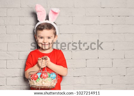 Cute little boy with bunny ears holding basket full of Easter eggs on brick wall background